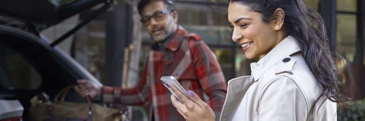 Man Loading Luggage into back of Vehicle and Woman Smiling at her Smartphone