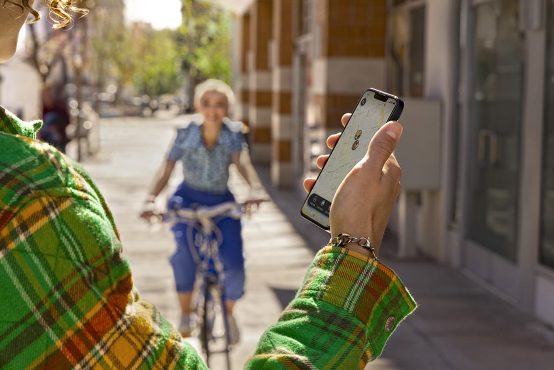 Woman in Distance Riding Bike Towards Man Holding Smartphone