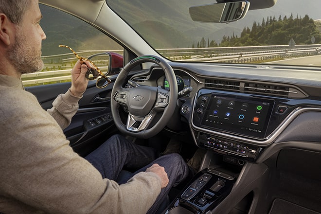 A person holds their eyeglasses in one hand and has their other hand on their knee while driving with Super Cruise driver assistance technology.