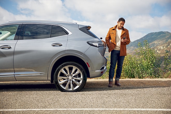 Man Stands at the Back of his Buick While Checking His Phone