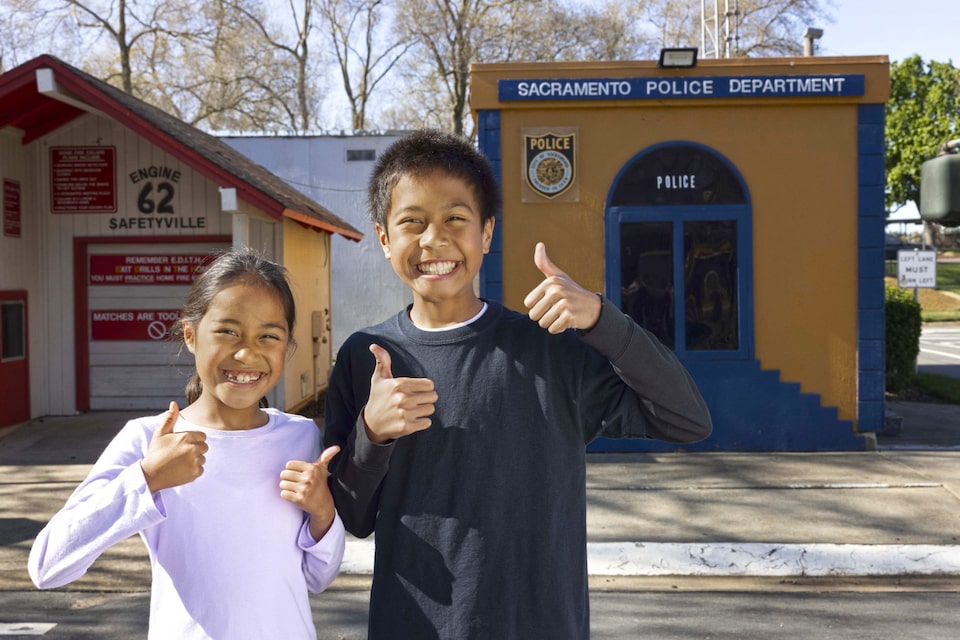 Two Kids Smiling and Giving Thumbs Up In Front of Buildings